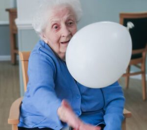 A resident from our Essex care home in the process of hitting a balloon during a seated exercise class. She is wearing a blue cardigan and has short white hair.