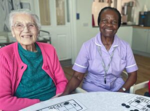 Abi sits with resident Nella. They are both smiling at the camera on International Women's Day. There are checker boards on the table.